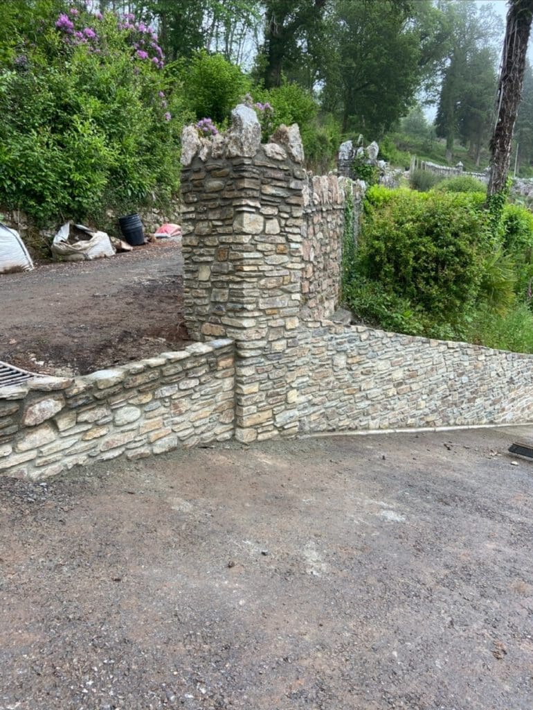 A stone wall with a decorative, castle-like pillar stands next to a gravel driveway—an inviting entrance often found near barn conversions—surrounded by greenery, trees, and blooming purple flowers.