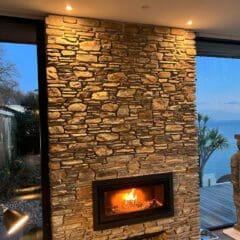 A modern living room with a stone masonry fireplace, fire burning inside. Large windows reveal an outdoor garden on the left and a sea view on the right. A statue stands by the window near the water—perfect inspiration for home renovations.