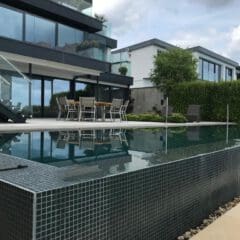 Modern house with large glass windows and balconies, featuring expert stone masonry accents, a patio with outdoor dining table and chairs, and an infinity-edge swimming pool in the foreground under a cloudy sky.