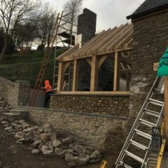 A person in an orange jacket stands near a stone building under construction, showcasing stone masonry work and exposed roof beams. Scaffolding, ladders, and materials typical of home renovations surround the site, with trees and a cloudy sky in the background.
