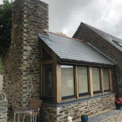 A stone house with a large chimney, slate roof, and corner windows with wooden frames showcases the charm of barn conversions. In front, there’s a small patio with a table, two chairs, and potted plants. The sky is overcast.