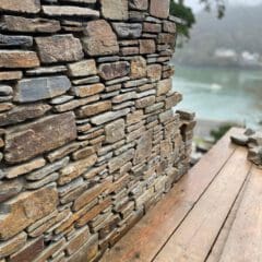 A close-up of a rustic stone wall under construction by skilled builders, with uneven stones stacked on top of each other; a weathered wooden surface is in the foreground and a blurred river and trees are visible in the background.