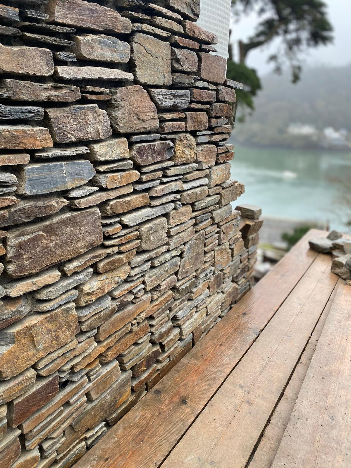 A close-up of a rustic stone wall under construction by skilled builders, with uneven stones stacked on top of each other; a weathered wooden surface is in the foreground and a blurred river and trees are visible in the background.