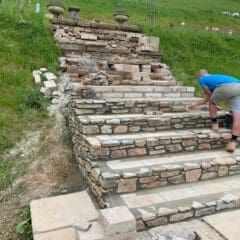 A person in a blue shirt and gray shorts is building wide stone steps on a grassy hillside, showcasing stone masonry skills. Partially finished steps and construction materials are visible.