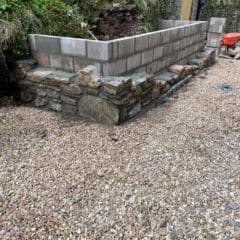 A partially constructed wall with a lower section of stone masonry and an upper section built from concrete blocks. The area around the wall is covered in gravel, with greenery and a stone building—ideal for barn conversions—in the background.