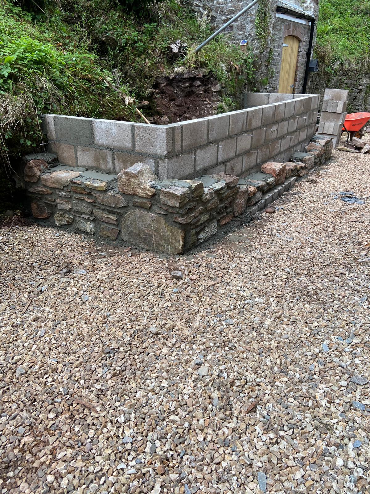 A partially constructed wall with a lower section of stone masonry and an upper section built from concrete blocks. The area around the wall is covered in gravel, with greenery and a stone building—ideal for barn conversions—in the background.