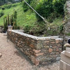A newly built stone retaining wall stands next to a gravel pathway, surrounded by greenery and shrubs on a sloping hillside—an ideal feature for home renovations or Passivhaus buildings. Construction tools and some cinder blocks are visible nearby.