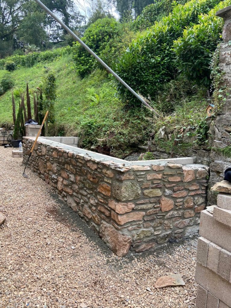 A newly built stone retaining wall stands next to a gravel pathway, surrounded by greenery and shrubs on a sloping hillside—an ideal feature for home renovations or Passivhaus buildings. Construction tools and some cinder blocks are visible nearby.