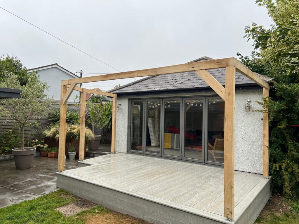 A wooden pergola frame stands on a light gray deck in front of a small modern garden building with large glass doors, surrounded by potted plants, lush greenery, and expert builders’ stone masonry on a cloudy day.