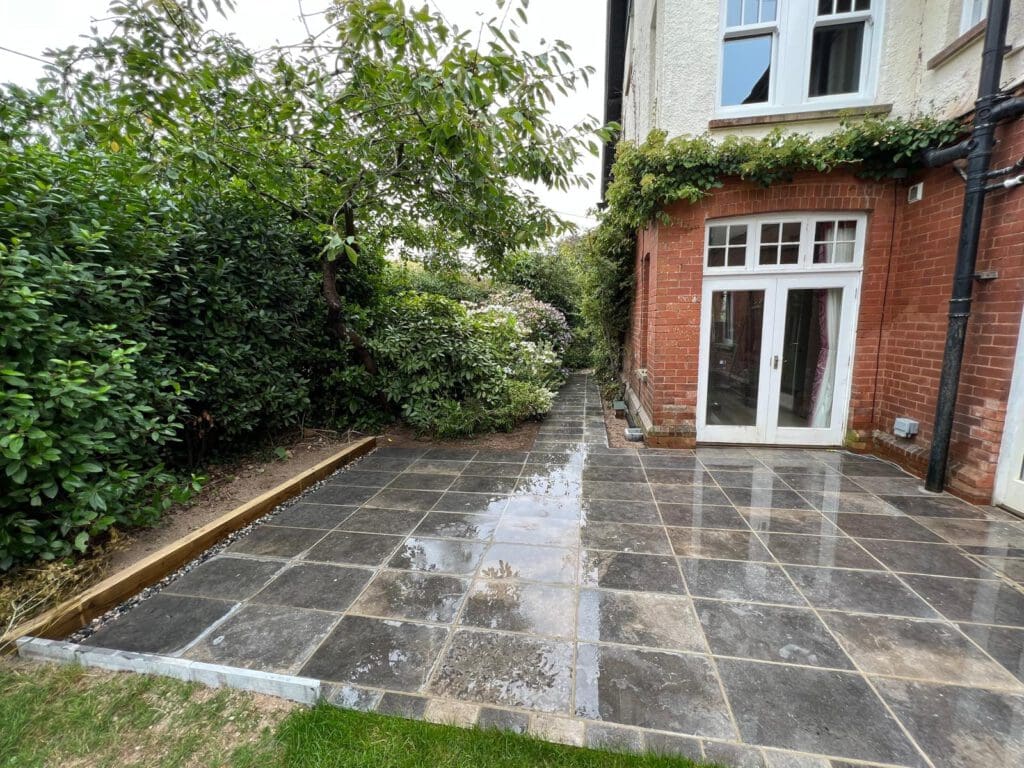 A newly laid patio with wet gray stone masonry tiles sits beside a red-brick and white house with French doors. Lush green bushes and a tree border the patio, and a narrow path leads alongside the house—perfect for home renovations.