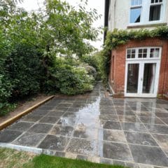 A newly laid patio with wet gray stone masonry tiles sits beside a red-brick and white house with French doors. Lush green bushes and a tree border the patio, and a narrow path leads alongside the house—perfect for home renovations.