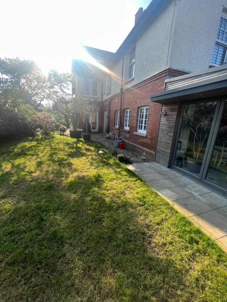 A sunlit garden with green grass is beside a two-story house of red brick and white walls. Built by skilled builders, sliding glass doors open to a patio, while shrubs line the garden's edge and sunbeams create bright highlights.