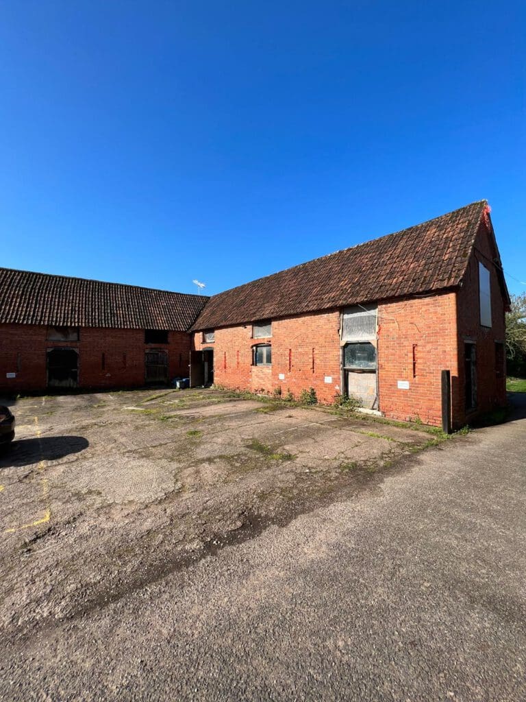 A large, old brick barn with a sloped tile roof stands under a clear blue sky. Perfect for barn conversions, it features several barred windows and doorways showing wear. The foreground is a paved, empty parking area.
