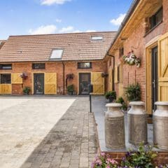 A courtyard with brick and stone masonry buildings featuring wooden doors and flower baskets, old-fashioned metal milk churns on a stone platform, and a paved walkway under a blue sky with scattered clouds.