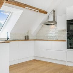 Modern attic kitchen with white cabinets, wooden countertops, and slanted ceiling with exposed beams. Large skylight lets in natural light. Expert builders crafted this space, featuring built-in oven, stove with hood, and light wood flooring.