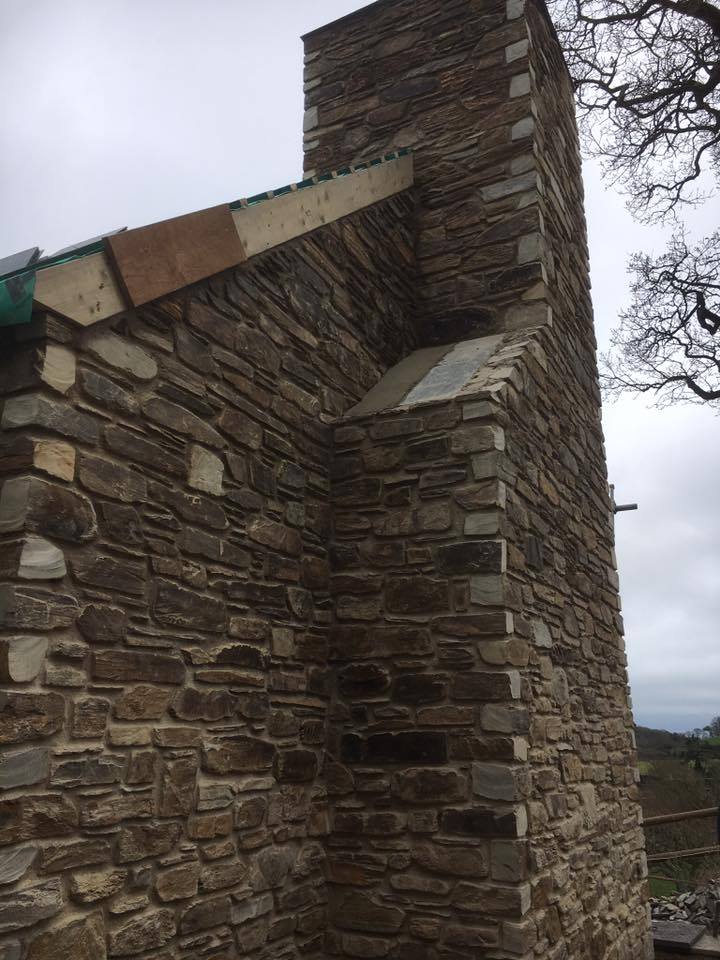 A close-up view of a tall stone chimney and wall under construction by skilled builders, with rough stones and temporary wooden boards at the roof edge. The sky is overcast, and a tree is partly visible to the right.
