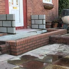 A partially built brick porch with two unfinished block pillars, a white front door, paving stones, a cement mixer, and builders' materials in front of a red brick house—perfect for home renovations.