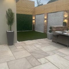 Modern backyard patio with large stone tiles, artificial grass, potted plants, and wicker dining furniture. Perfect for home renovations, this cozy space features wall lights and a wooden privacy fence for warm evenings outdoors.
