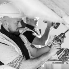 Two builders wearing hard hats and safety vests use power drills to work on a ceiling, possibly during barn conversions. One is in the foreground and the other in the background. The image is in black and white.