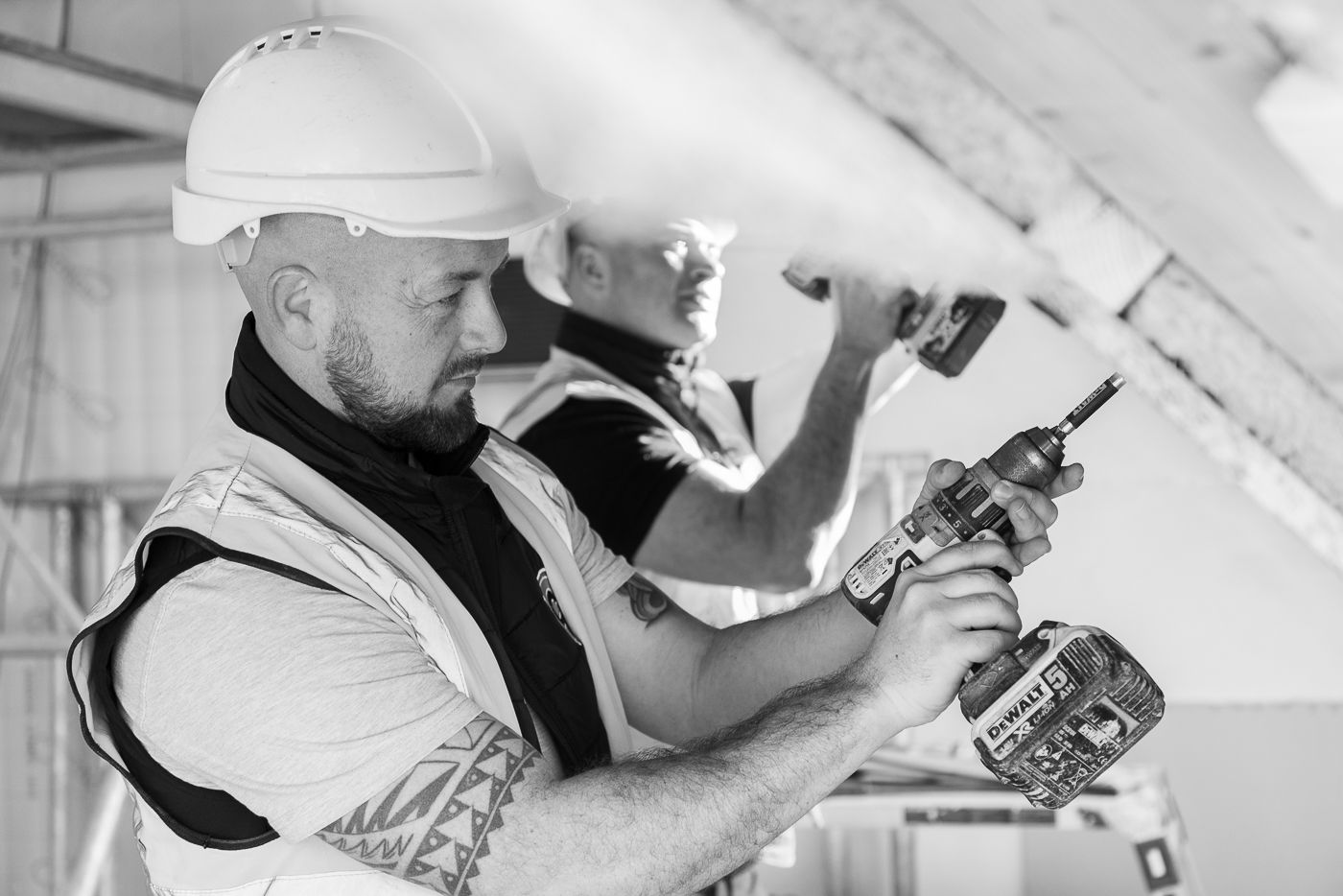 Two builders wearing hard hats and safety vests use power drills to work on a ceiling, possibly during barn conversions. One is in the foreground and the other in the background. The image is in black and white.