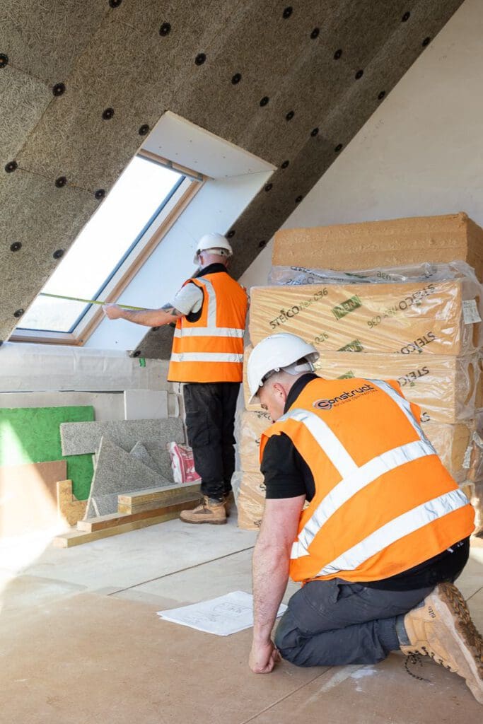 Two builders in safety vests and helmets work in an attic; one kneels over plans while the other measures a skylight window, preparing for energy-efficient Passivhaus buildings. Insulation materials are stacked nearby.