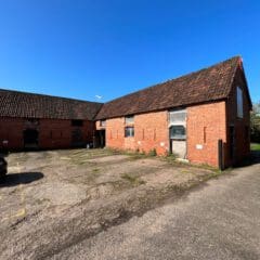 A large brick barn with a tiled roof, weathered doors, and boarded windows stands under a clear blue sky. Crafted by skilled builders using traditional stone masonry, it sits on paved ground with a small parking area visible to the left.